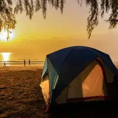 Wschód słońca nad morzem, gdzie rozbito luksusowe pole namiotowe. Ludzie spacerują po plaży, a namioty kuszą komfortem.