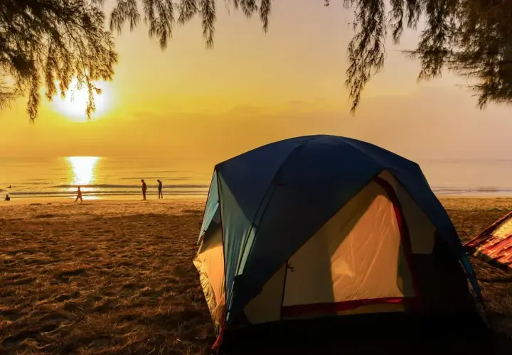 Wschód słońca nad morzem, gdzie rozbito luksusowe pole namiotowe. Ludzie spacerują po plaży, a namioty kuszą komfortem.