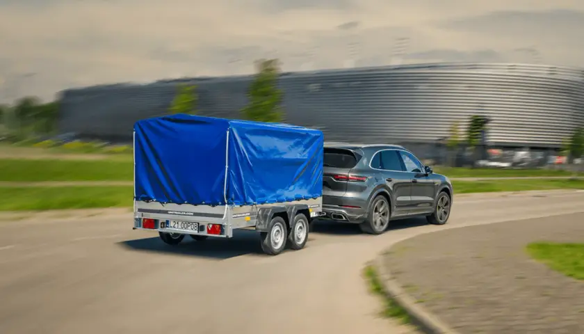 Szary SUV z przyczepą pod niebieską plandeką jedzie z dużą prędkością, mijając nowoczesny budynek.