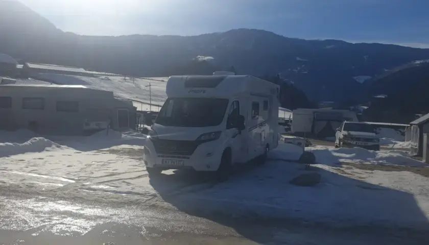 Kampery i przyczepy kempingowe na śniegu, gotowe na całoroczny camping w górach. Słońce przebija się przez chmury, oświetlając zimowy krajobraz.