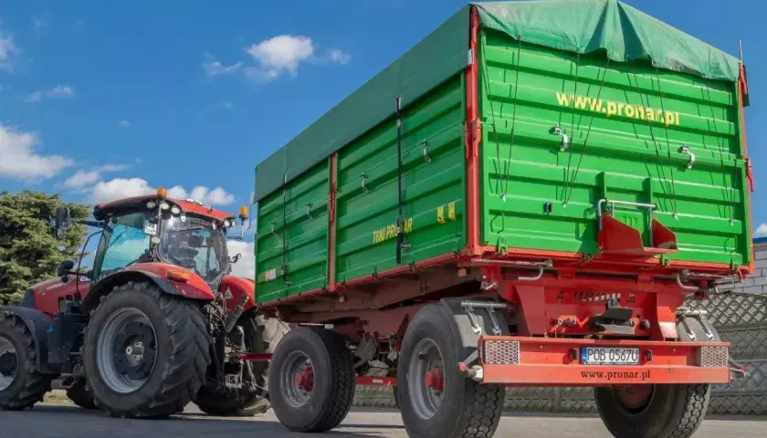 Czerwony traktor ciągnie zieloną przyczepę z plandeką. Duża masa przyczepy gotowa do pracy na polu.