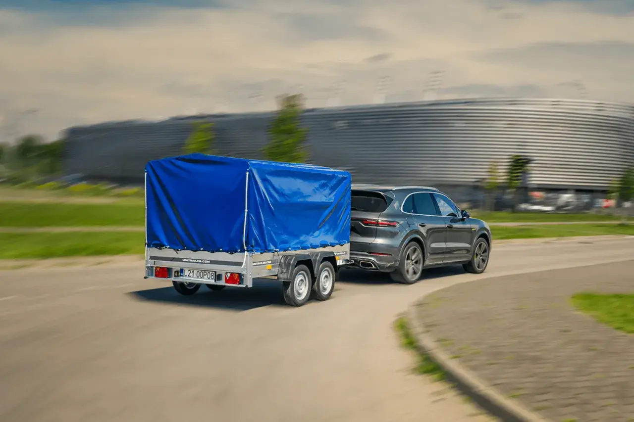 Szary SUV z przyczepą pod niebieską plandeką jedzie z dużą prędkością, mijając nowoczesny budynek.