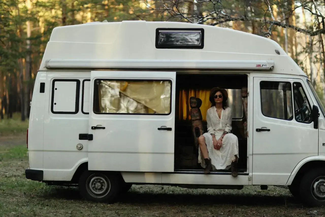 Kobieta w białej sukience siedzi w otwartych drzwiach białego kampera, otoczona lasem. Idealne polskie caravaning miejscówki na wakacje.