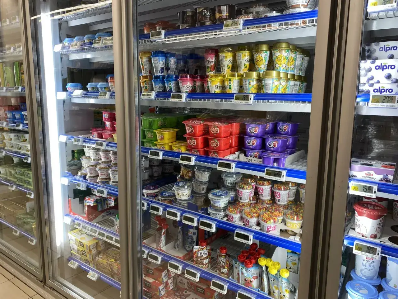 W lodówce w Norwegii widać jogurty i desery. Ceny w sklepach są widoczne na etykietach, np. 27,90 za jogurt.