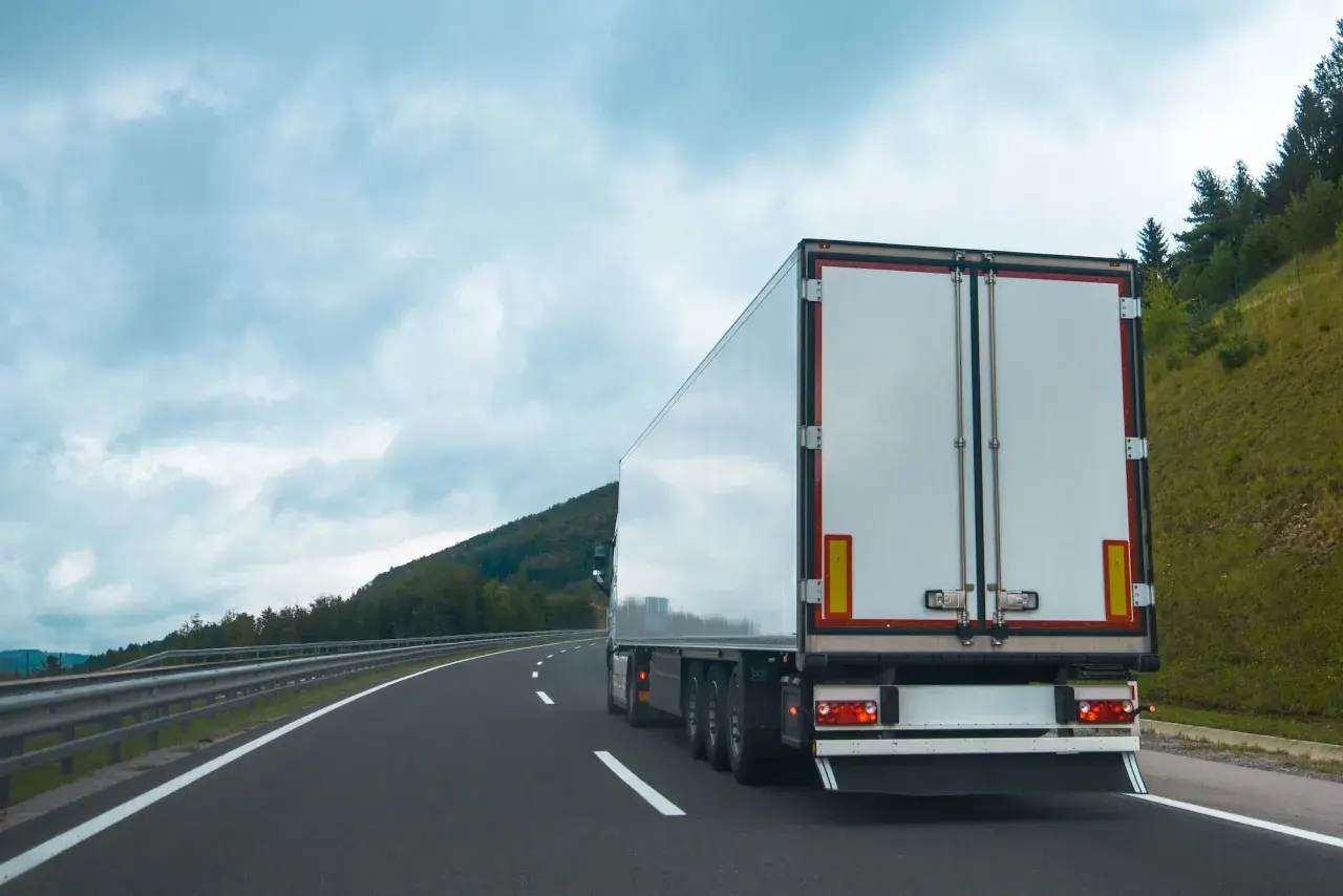 Ciężarówka na autostradzie, z tyłu widać białą naczepę. Na poboczu rosną drzewa, a nad nimi chmury. Tablice wyróżniające przepisy są widoczne na naczepie.