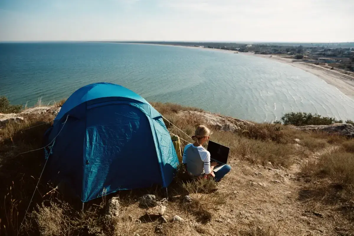 Niebieski namiot i osoba z laptopem na wzgórzu, z widokiem na spokojne, lazurowe morze. Idealny camping lazurowe wybrzeże.