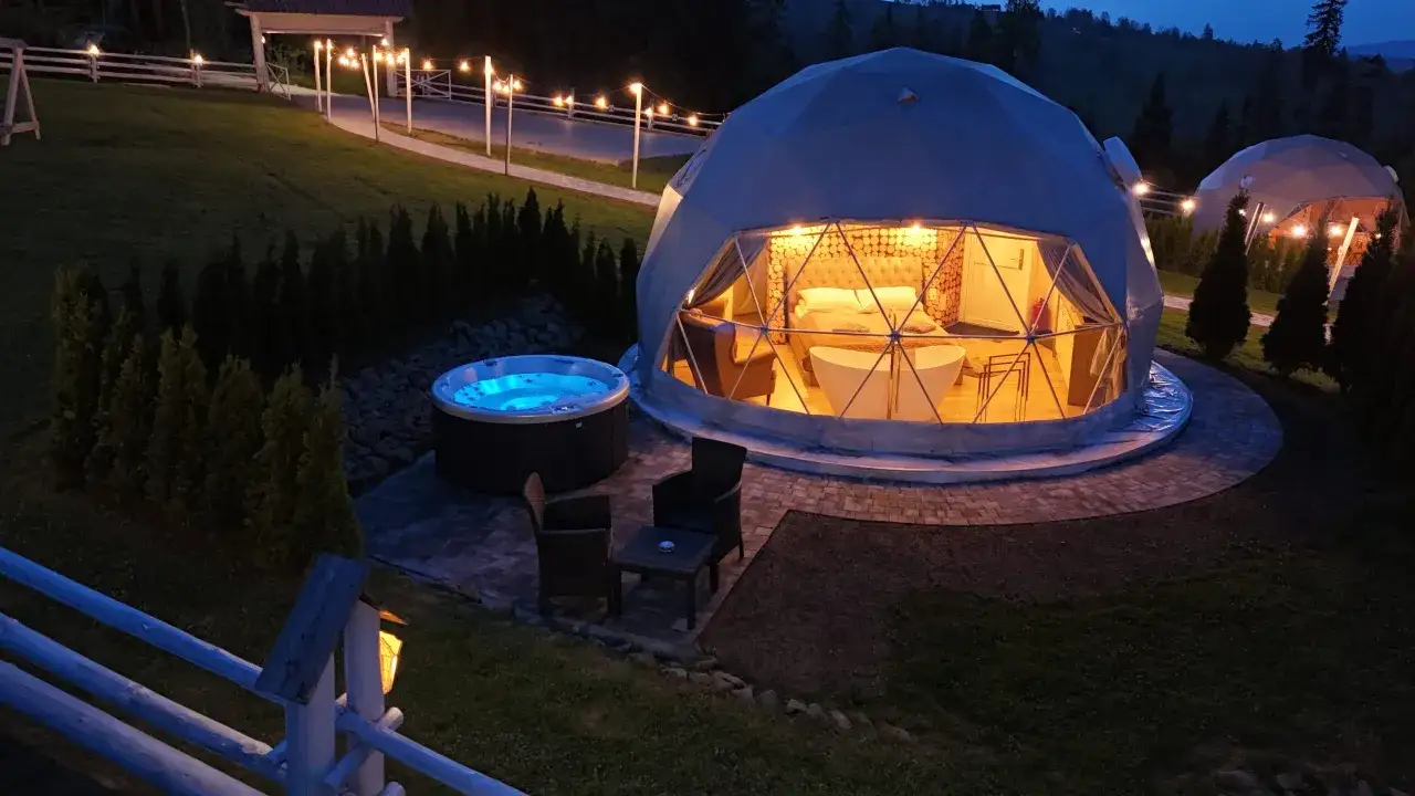 Glamping co to? Luksusowy namiot kopułowy z wanną i łóżkiem, obok jacuzzi i mebli ogrodowych, oświetlony girlandami.