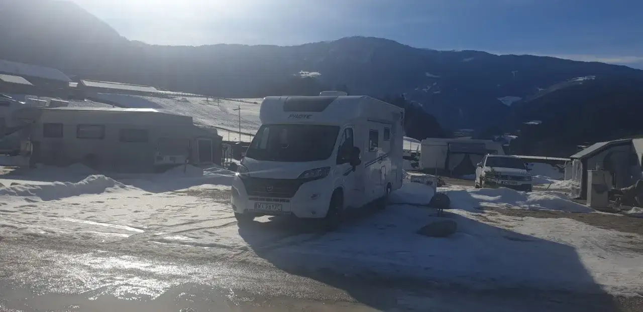 Kampery i przyczepy kempingowe na zaśnieżonym polu namiotowym. Słońce przebija się przez góry, tworząc idealne warunki do całorocznego campingu.