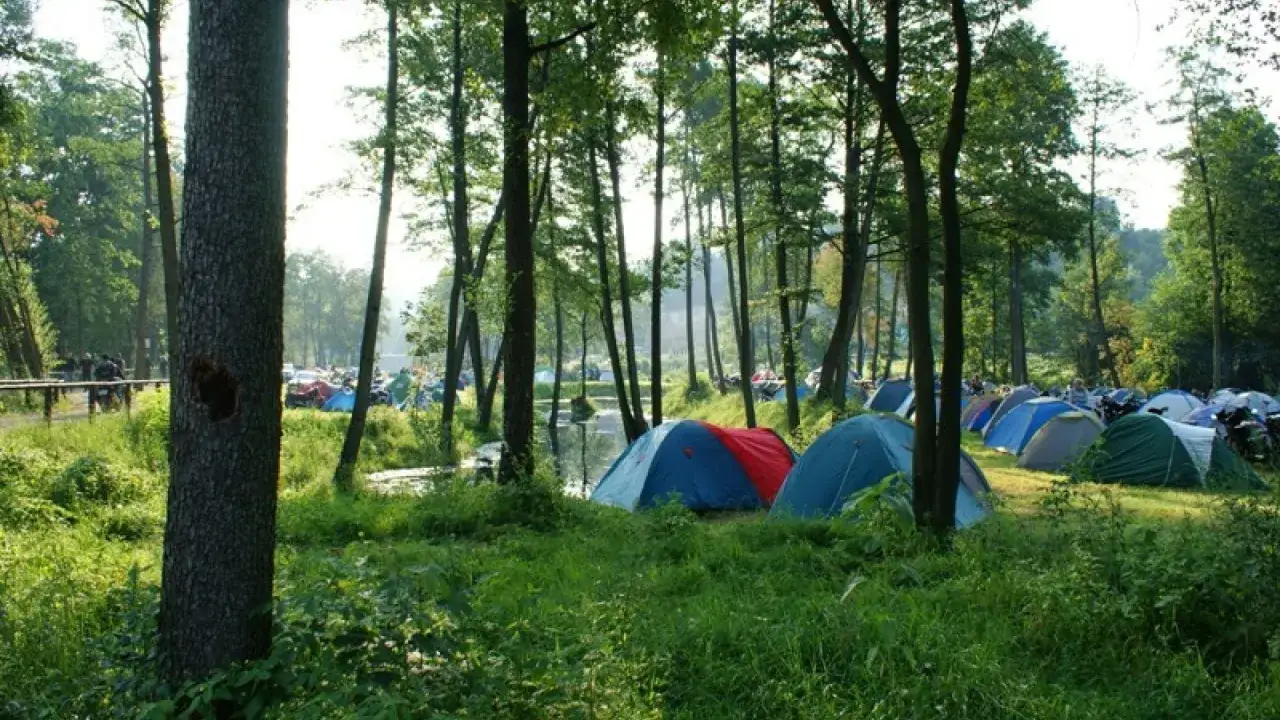 Kolorowe namioty rozstawione w lesie, nad wodą. W tle widać motocykle i ludzi. To idealne miejsce na camping Świętokrzyskie.