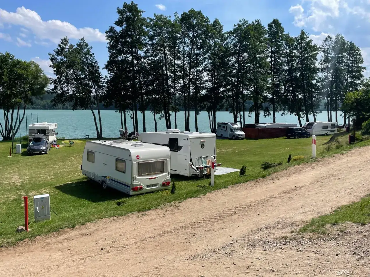 Słoneczny dzień na camp Polska nad jeziorem. Przyczepy kempingowe i kampery na zielonej trawie, w tle las i błękitna woda.