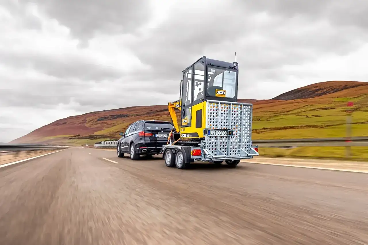 Czarne BMW ciągnie przyczepę z minikoparką JCB. Czy można ciągnąć przyczepę dwuosiową na kat. B?